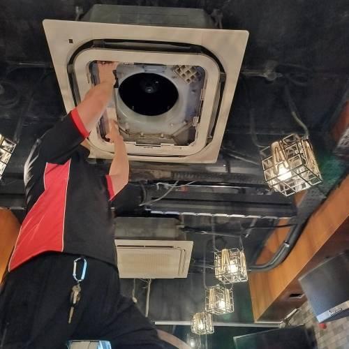 Expert Ceiling Aircon Maintenance: Ensuring Your Unit’s Top Performance A skilled technician attentively repairs or inspects an air conditioning unit installed on the ceiling, amidst an industrial setting with dark ceilings and protective wire-caged light fixtures.