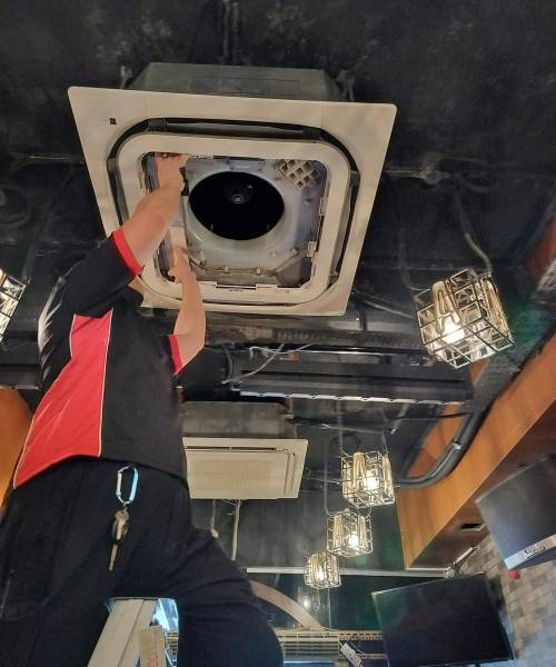 Expert Ceiling Aircon Maintenance: Ensuring Your Unit’s Top Performance A skilled technician attentively repairs or inspects an air conditioning unit installed on the ceiling, amidst an industrial setting with dark ceilings and protective wire-caged light fixtures.