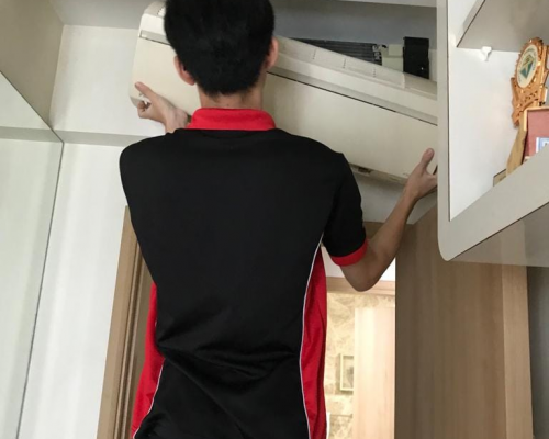 Professional Aircon Servicing: Efficient Maintenance by a Skilled Technician A technician in black and red attire servicing a white air conditioning unit installed near the ceiling in a room with light-colored walls and wooden cabinets.