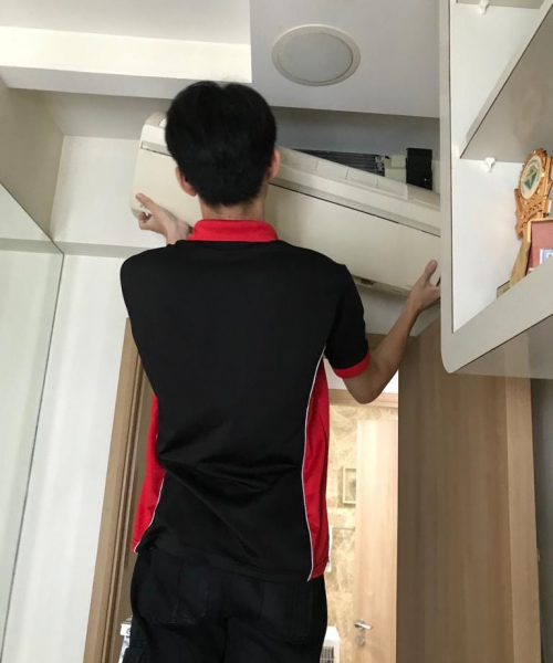 Professional Aircon Servicing: Efficient Maintenance by a Skilled Technician A technician in black and red attire servicing a white air conditioning unit installed near the ceiling in a room with light-colored walls and wooden cabinets.